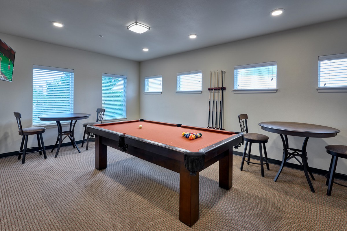 A cozy game room featuring a pool table with a set of billiard balls arranged, surrounded by bar stools and windows for natural light.