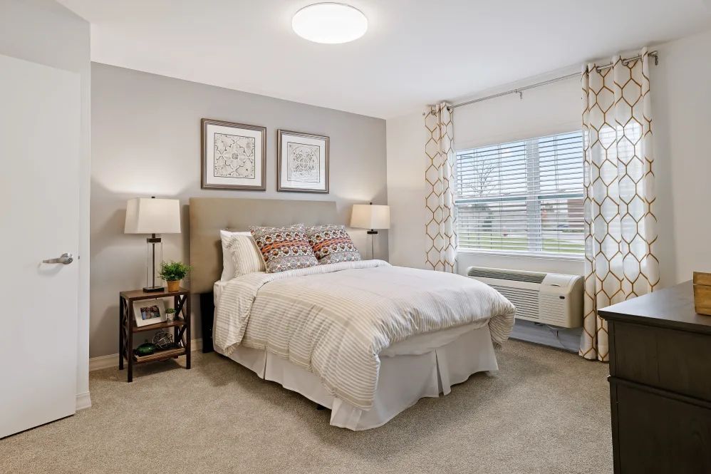 A modern bedroom with neutral tones, featuring a comfortable bed, lamps, and large windows.