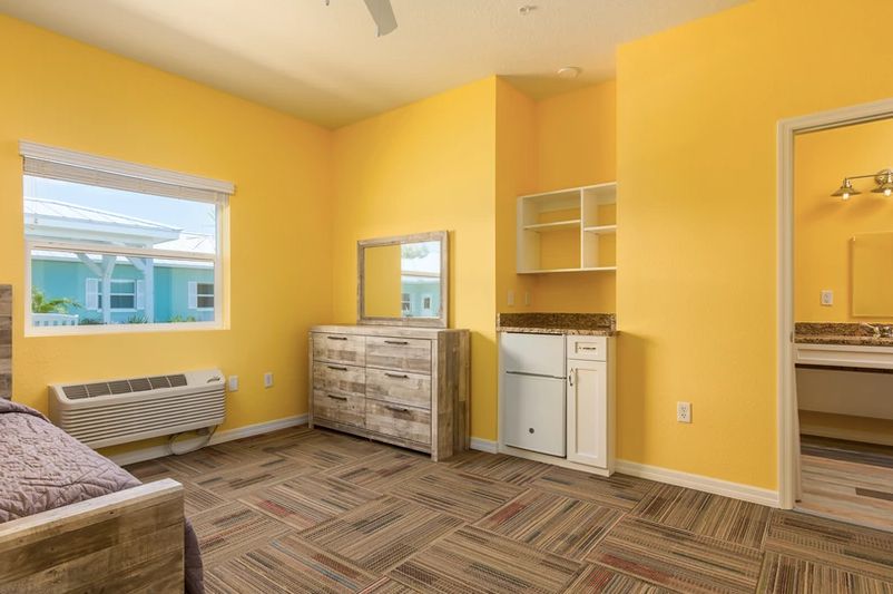 A brightly lit room with yellow walls, featuring a dresser, window, and open shelves.