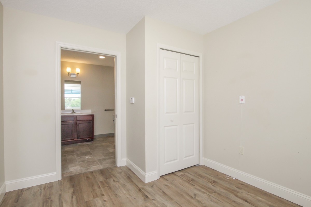 An empty bedroom with a view of a bathroom through the doorway, featuring hardwood floors and neutral-colored walls.