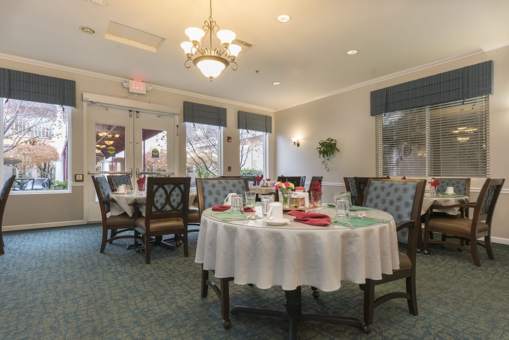 An elegant dining room with tables set for a meal, featuring a chandelier and large windows.