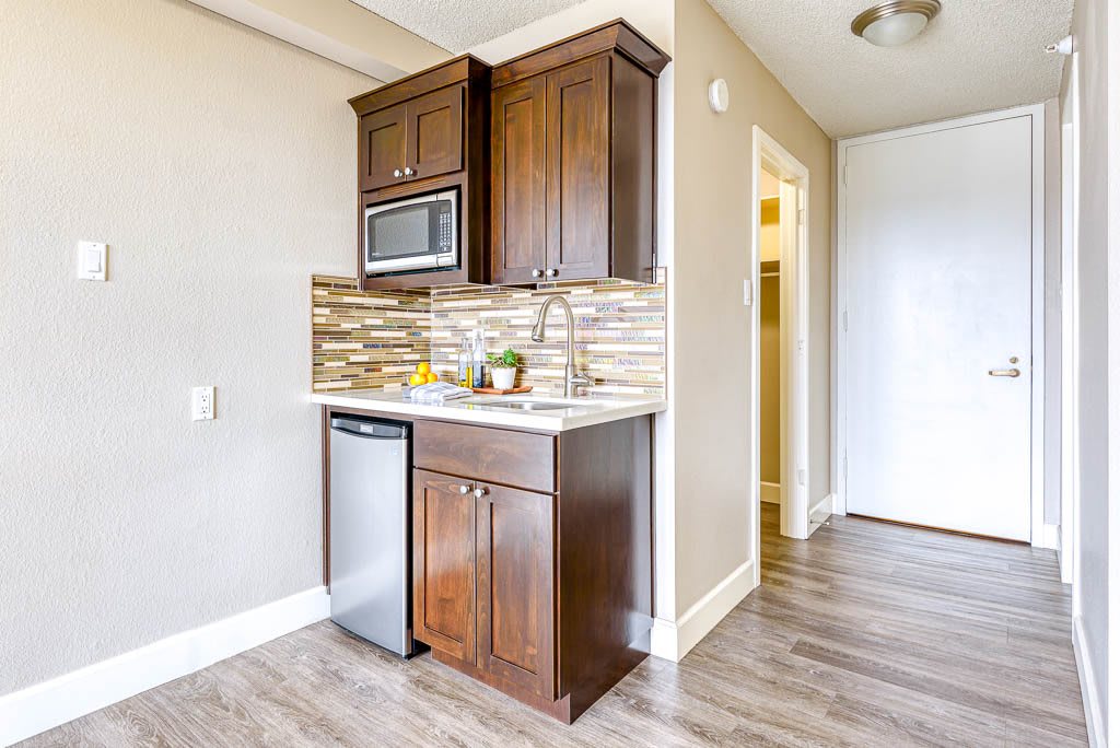 A compact kitchenette with wooden cabinetry, a mini fridge, and a microwave set against wood flooring.