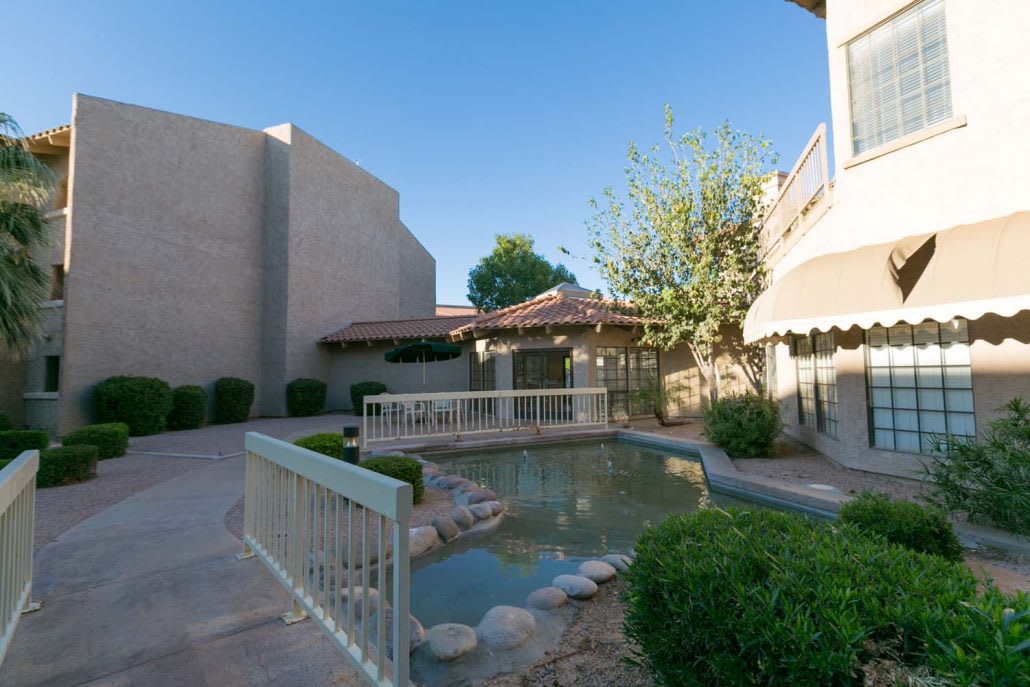 A tranquil courtyard featuring a small pond with a bridge, surrounded by greenery and buildings under a clear blue sky.