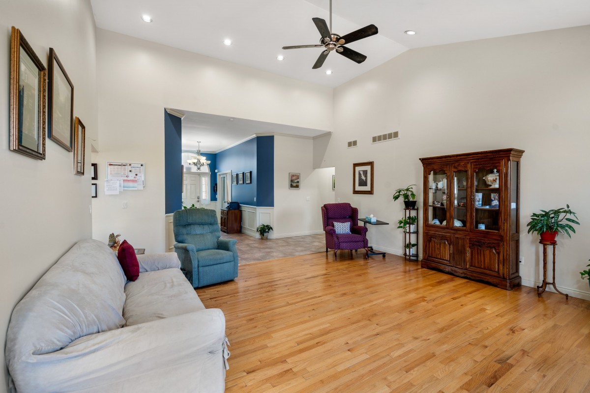 A spacious living room with wooden flooring, featuring a couch, armchairs, ceiling fan, and a display cabinet.