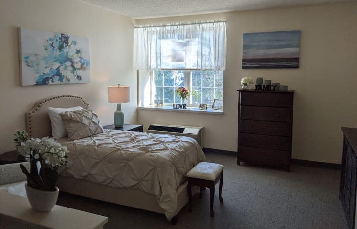A cozy bedroom with a neatly made bed, artwork on the walls, and light streaming in through a window.
