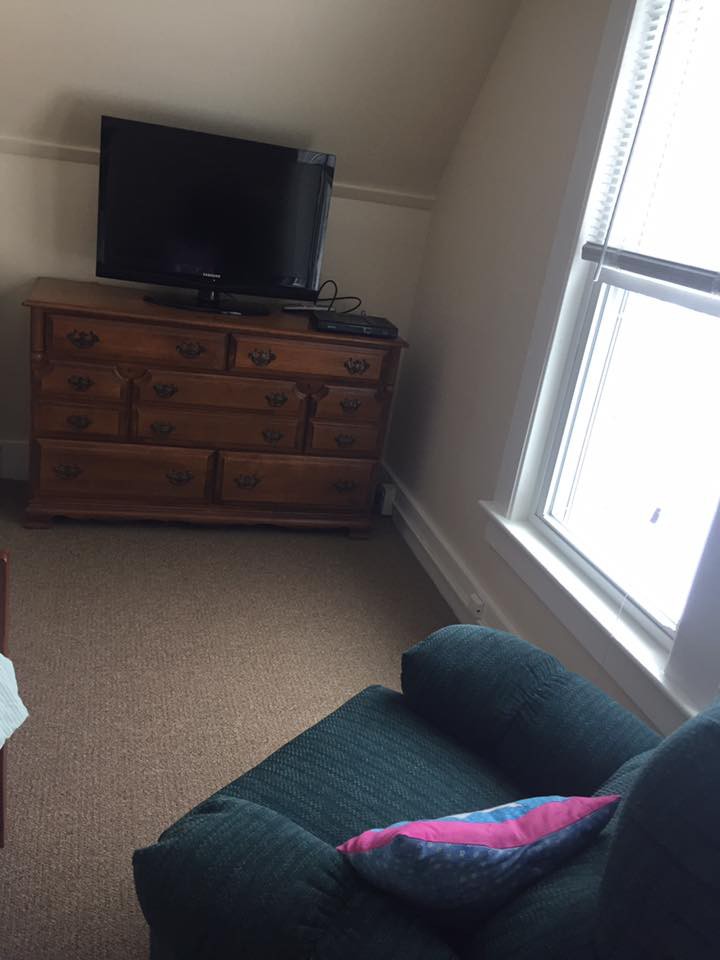 A cozy living room corner with a TV on a dresser, next to a comfortable chair and a large window.