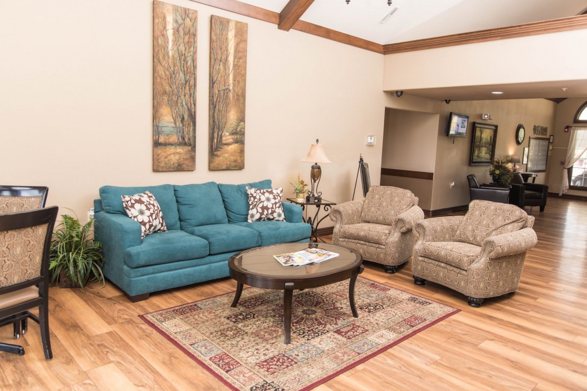 A cozy living room featuring a blue sofa, two beige armchairs, and a wooden coffee table atop a patterned rug. The room is decorated with wall art and has hardwood flooring.