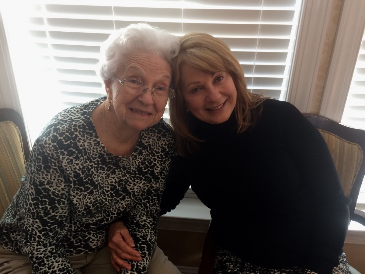 An elderly woman and a younger woman sitting together and smiling indoors.