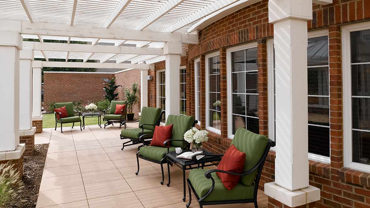 A stylish patio area with comfortable seating under a pergola, featuring green cushions and red pillows against a brick wall.