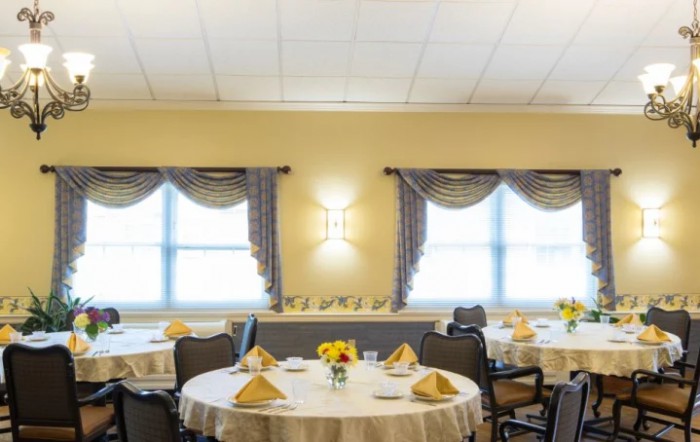 A well-decorated dining room featuring round tables with neatly arranged table settings, set beside large windows with draped curtains.