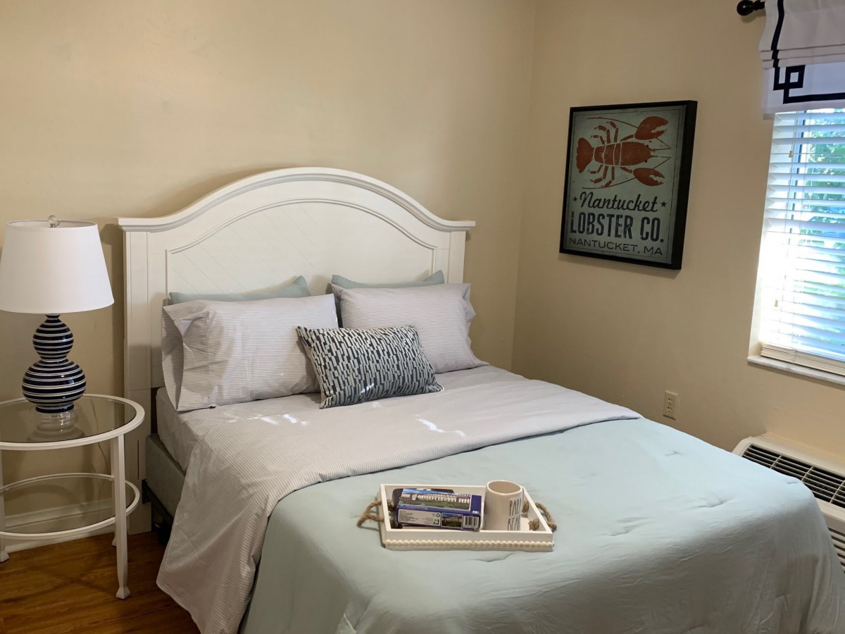 A cozy and neatly arranged bedroom with a white bed, decorative pillows, a bedside lamp, and framed artwork on the wall.