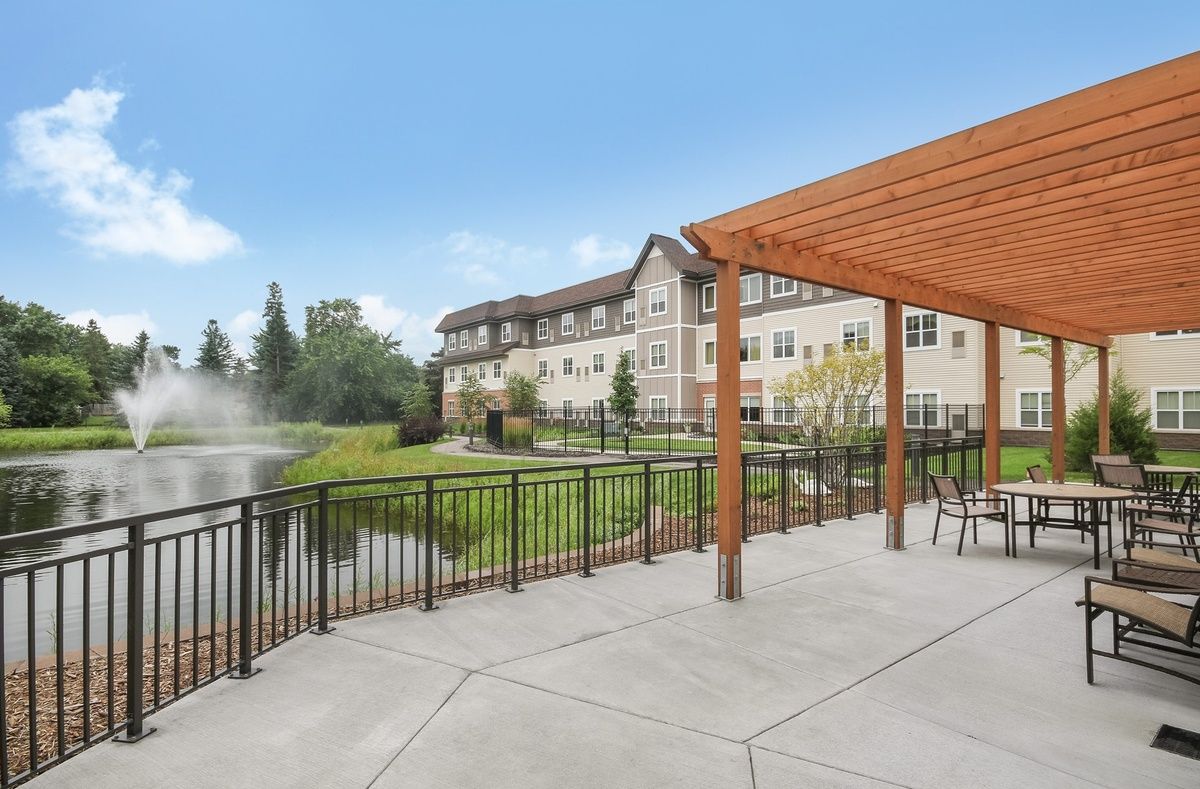 A scenic view of a building near a pond, with an outdoor seating area under a wooden pergola.