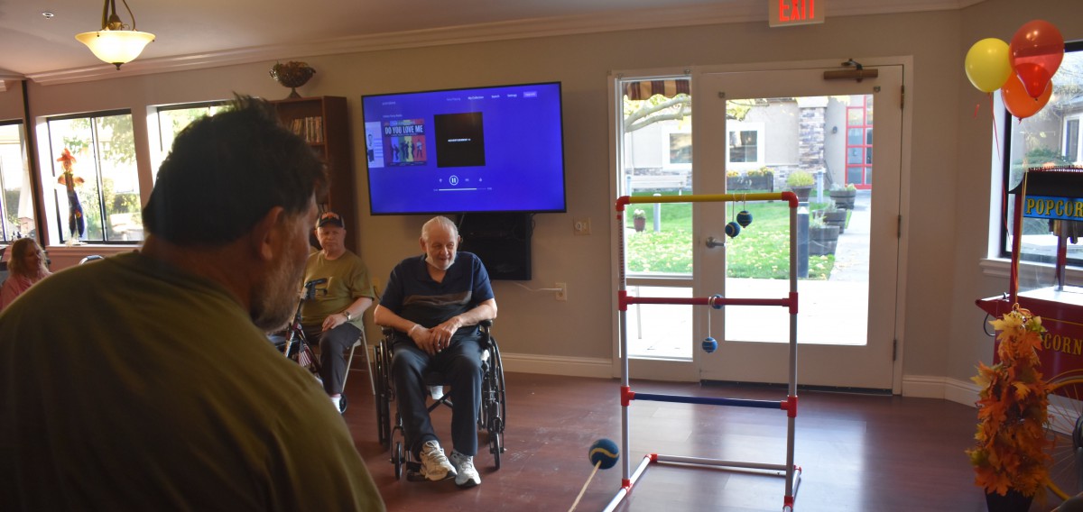 A group of people gather indoors, with one person in a wheelchair near a ladder toss game setup.