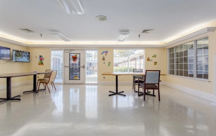 An interior room with tables, chairs, and large windows providing natural light.