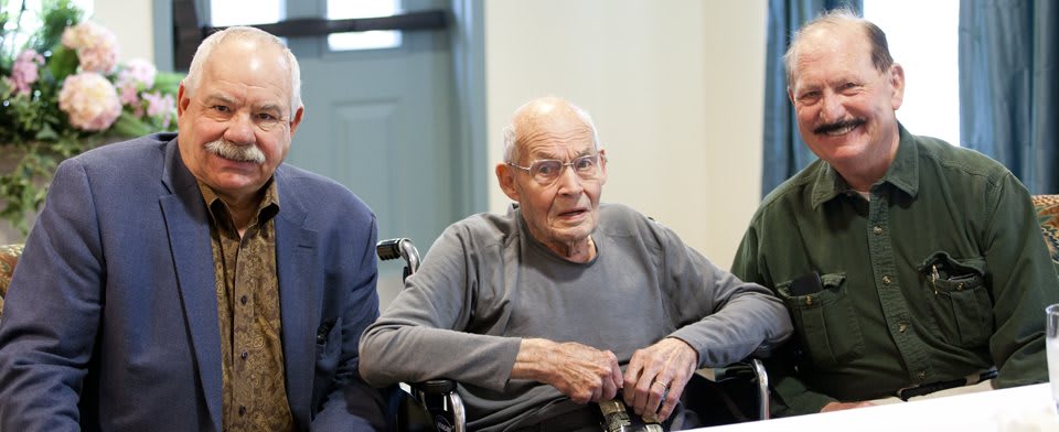 An elderly man wearing glasses is seated in a wheelchair indoors.
