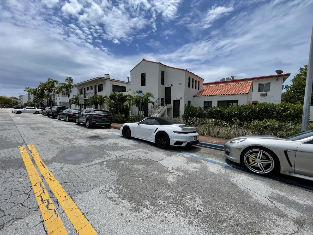 A row of parked cars in front of residential buildings under a cloudy sky.