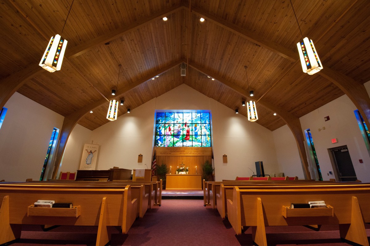 Interior view of a church with wooden pews and a stained glass window at the front.