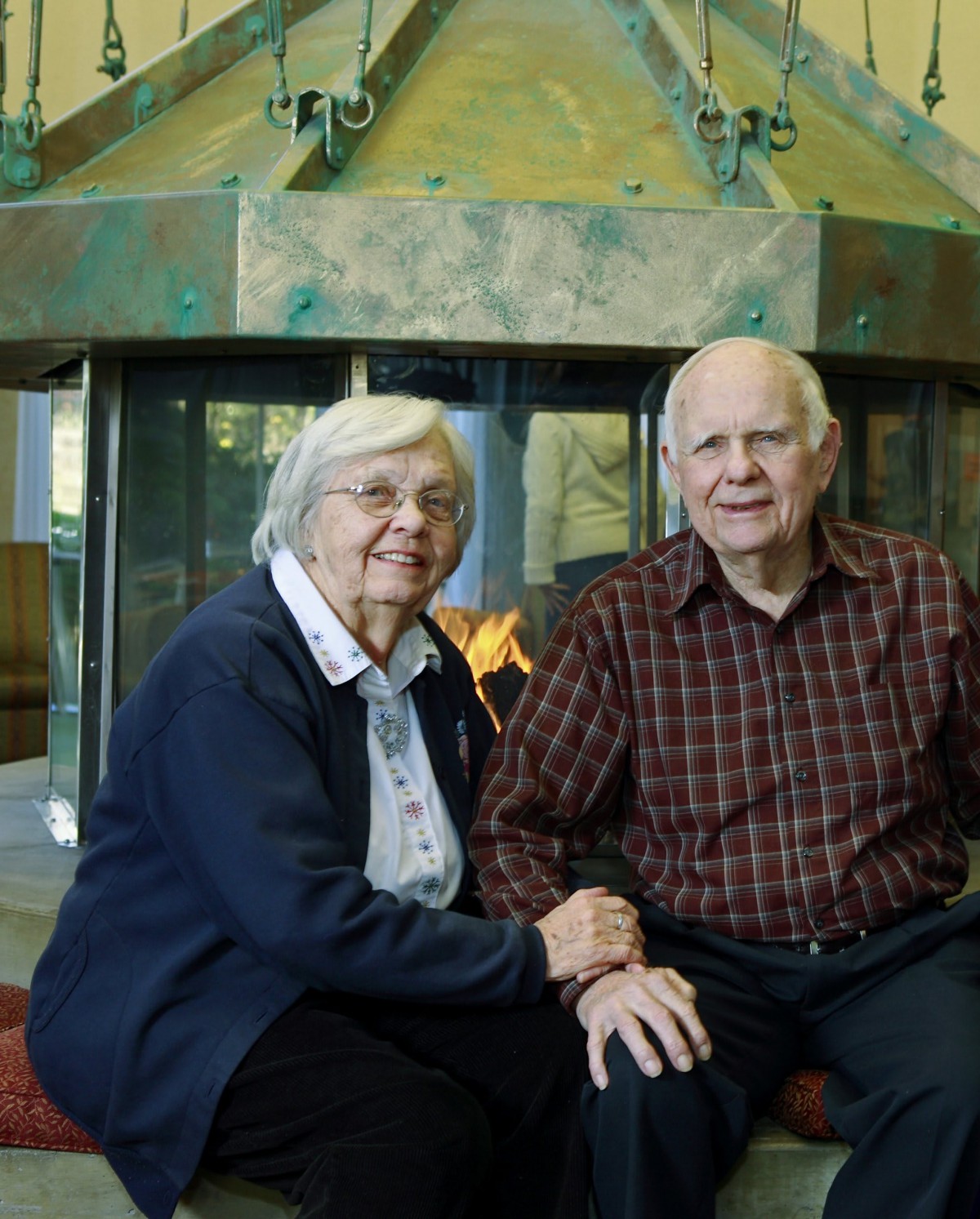 An elderly couple sitting together in front of a fireplace.