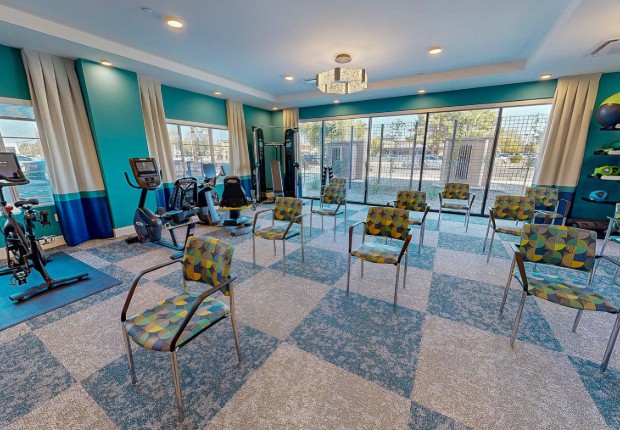 A spacious exercise room with arranged chairs for group activities, featuring gym equipment and large windows for natural lighting.