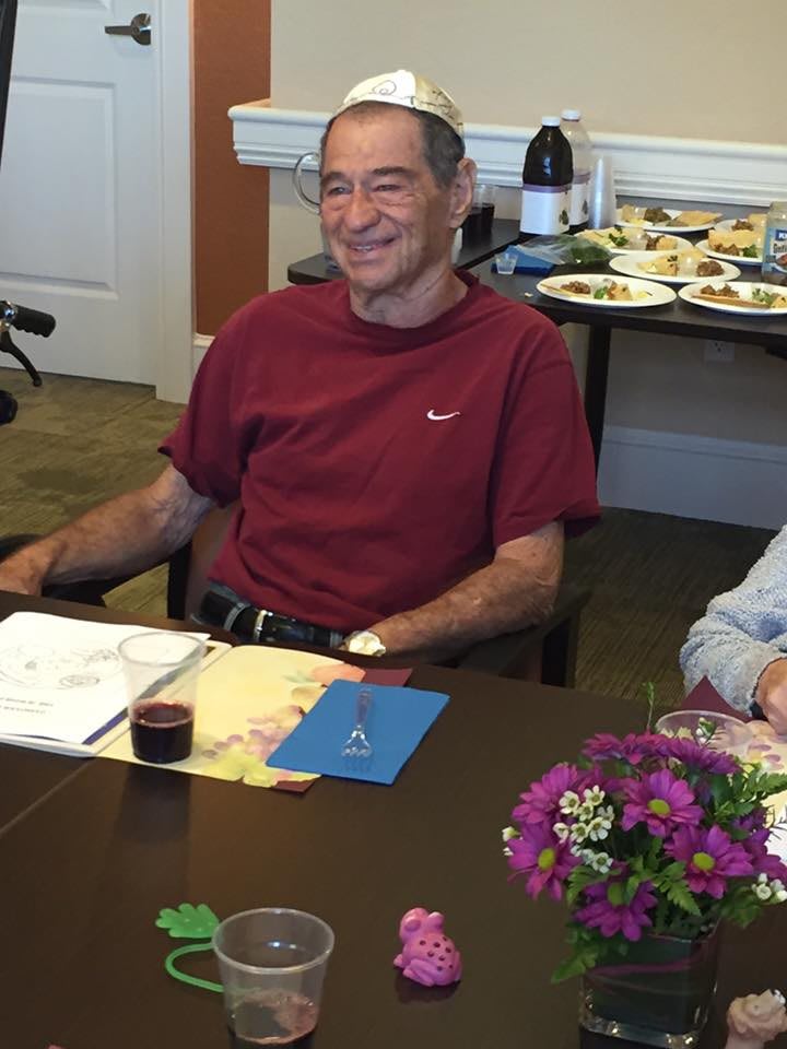 A man in a red shirt is smiling while sitting at a table indoors, with food and flowers around.