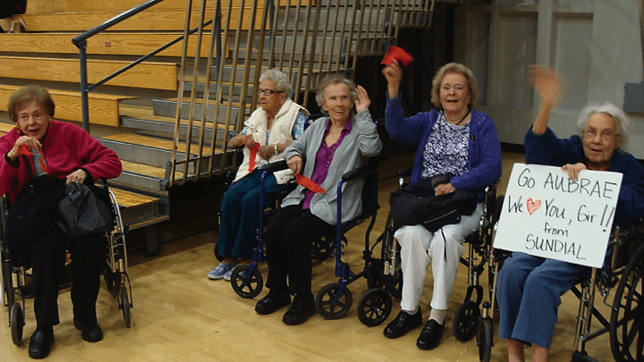 A group of elderly women sitting in wheelchairs indoors, smiling and waving red objects.