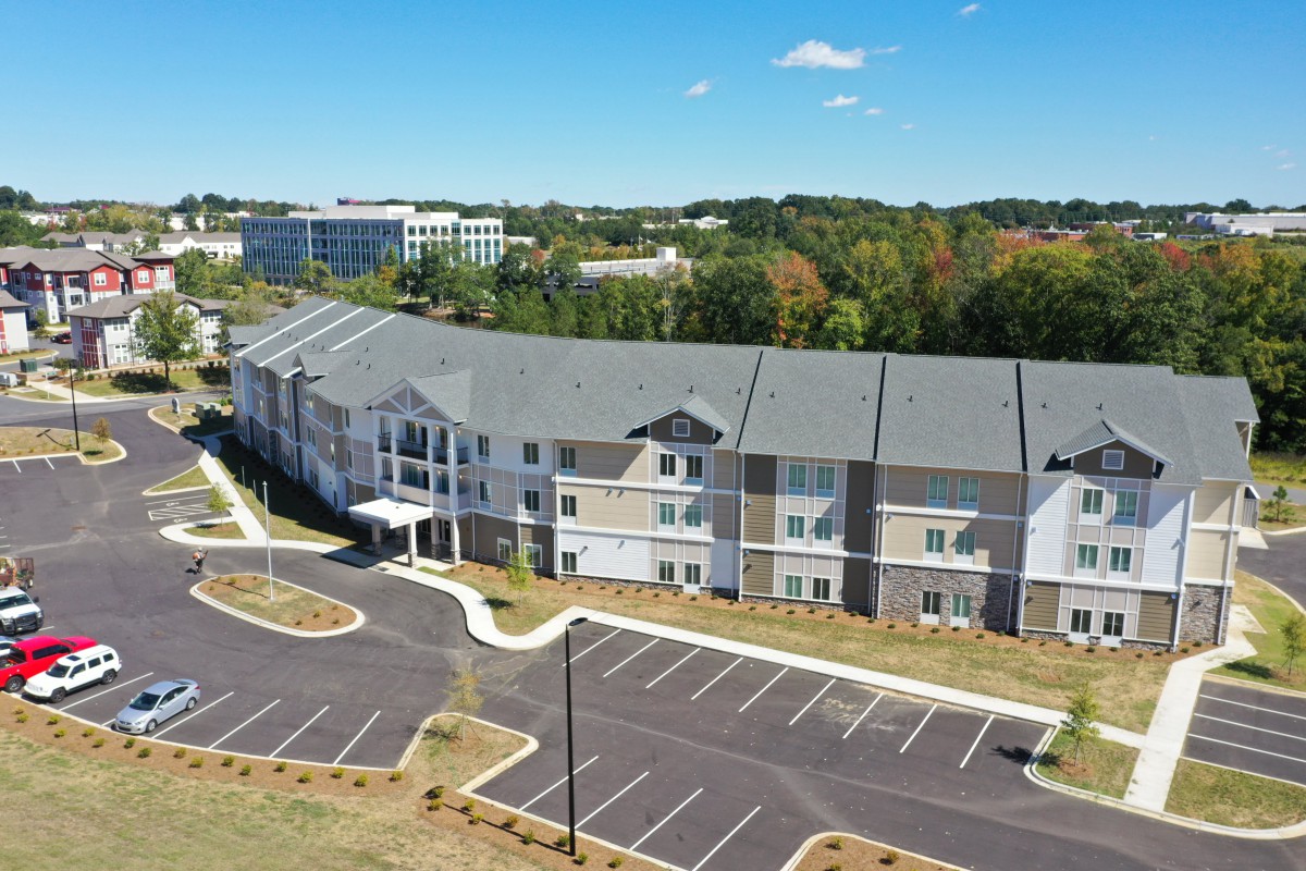 An aerial view of a modern residential apartment complex with an adjacent parking lot, surrounded by trees and other buildings in the background.