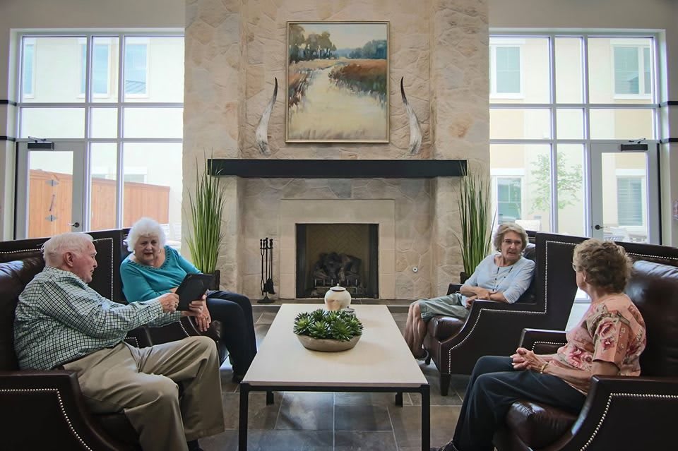 A group of seniors sitting in a cozy living room, engaged in conversation near a fireplace.