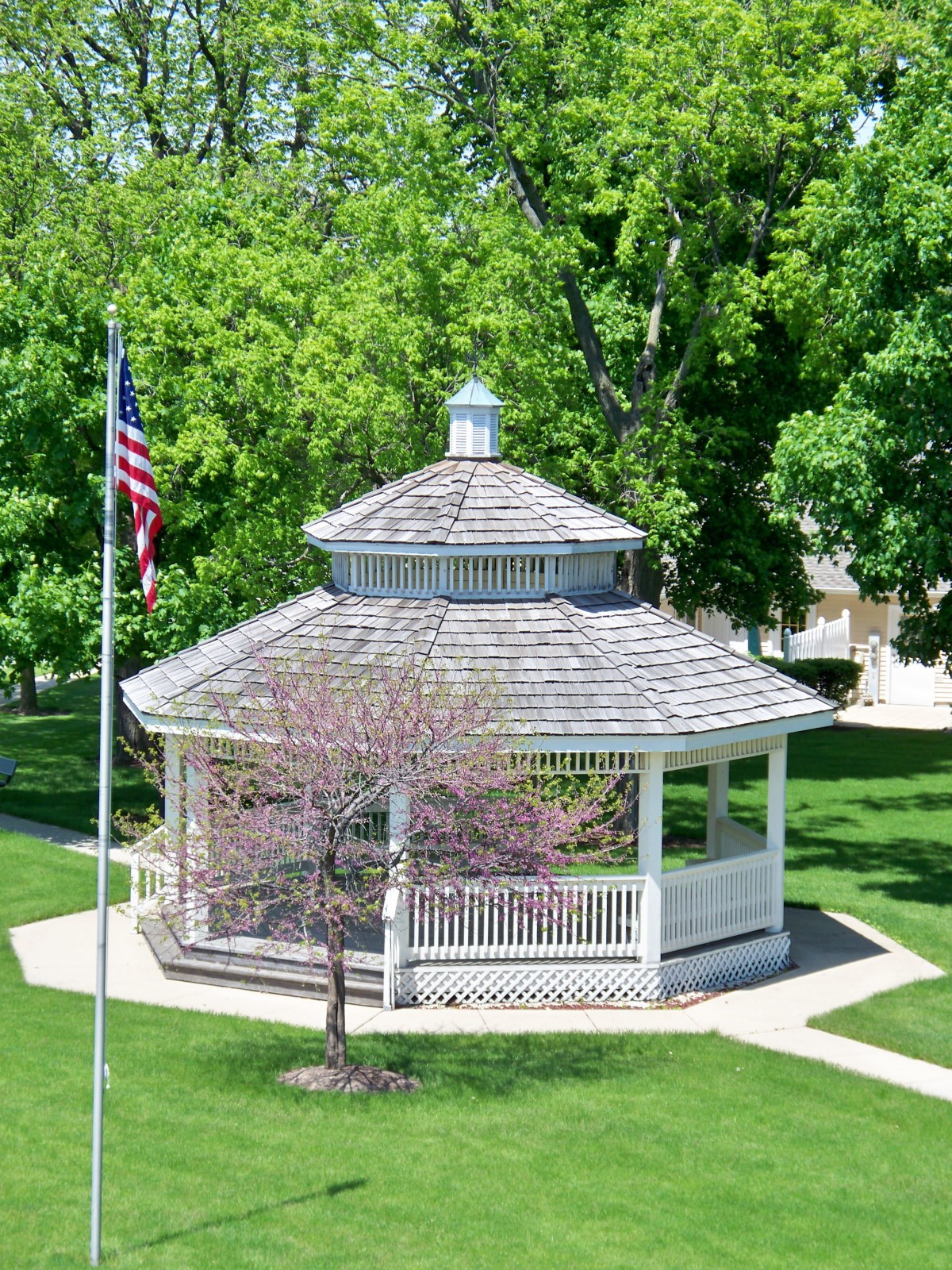 A charming gazebo in a park setting with a flagpole and lush green trees.