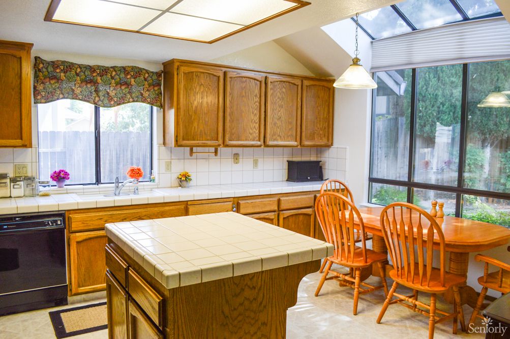 A bright kitchen with wooden cabinets, a tiled island, and orange dining chairs under ample natural light.