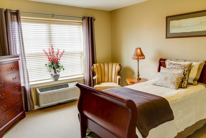 A cozy bedroom featuring a wooden bed, chair, and a window with flower decor.