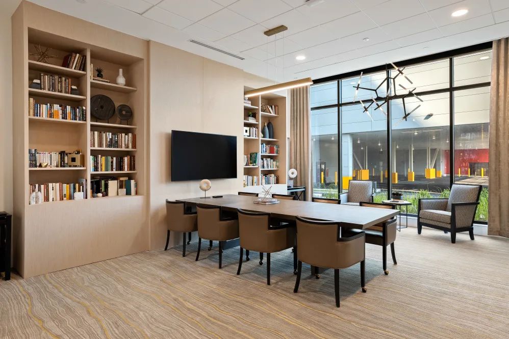 A modern conference room with a large table, comfortable chairs, and bookshelves, featuring a television and large windows for natural light.