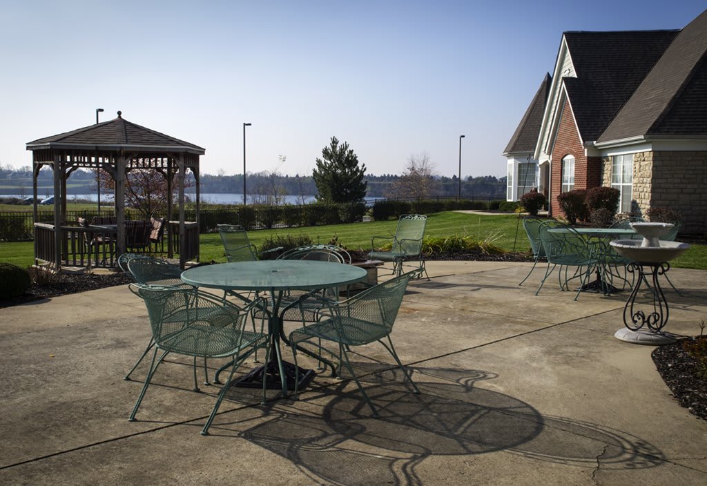 A sunny patio area featuring metal tables and chairs, with a gazebo and a lake in the background.
