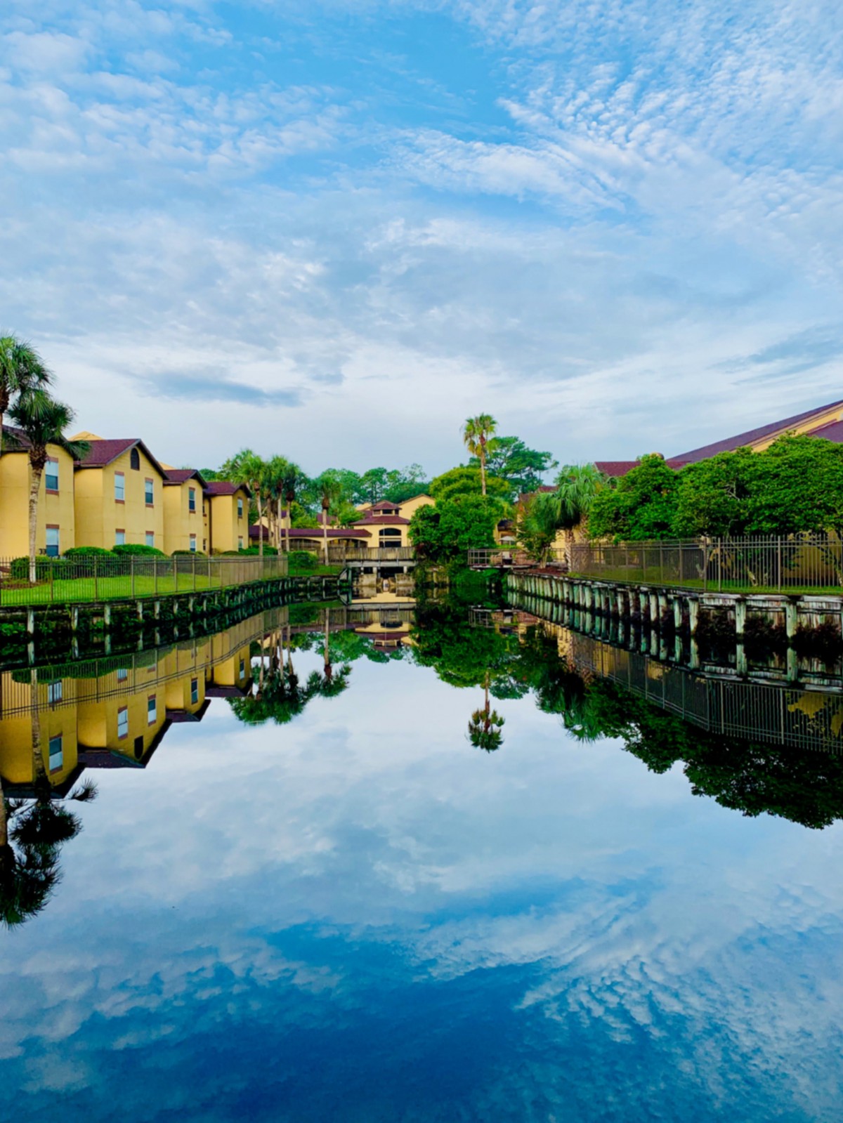 A serene view of buildings and trees reflected in a calm river under a blue sky.
