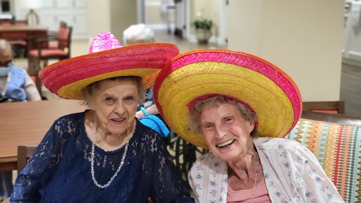 Two elderly women smiling and wearing colorful sombreros indoors.