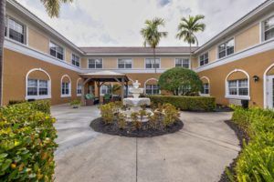 A courtyard with a central fountain, surrounded by lush landscaping and a two-story building with palm trees.