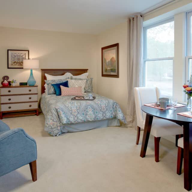 A cozy bedroom with elegant decor, featuring a bed, a small dining table with chairs, and a dresser with a lamp.