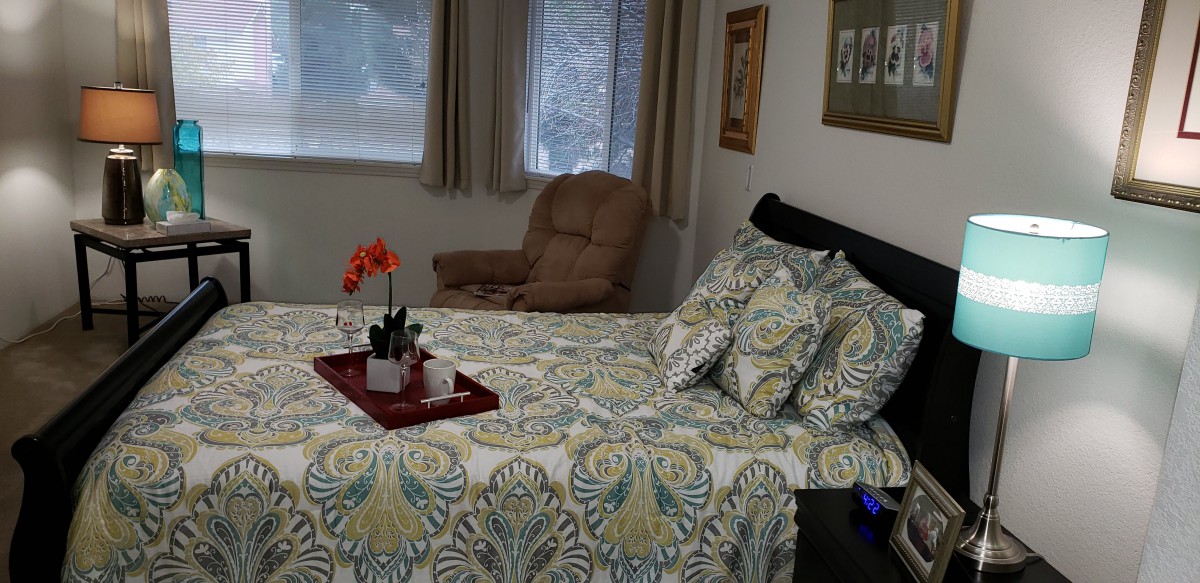 A cozy bedroom featuring patterned bedding, a tray on the bed, and a comfortable chair by the window.