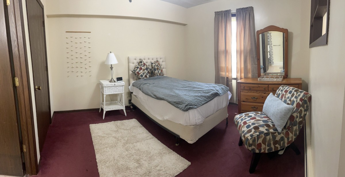 A cozy bedroom with a single bed, nightstand, and lamp, featuring warm curtains and carpeting.