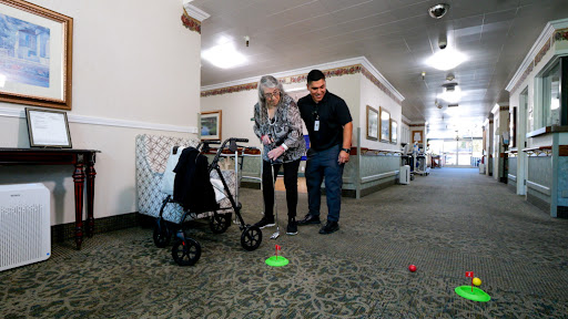 An elderly woman participates in a physical therapy activity with the assistance of a caregiver in a hallway setting.