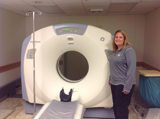A woman stands next to a CT scanner in a healthcare setting.