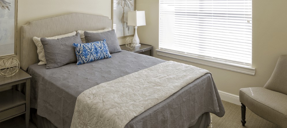 A cozy bedroom with a neatly made bed, featuring gray and blue pillows, a bedside table with a lamp, and natural light from the window.
