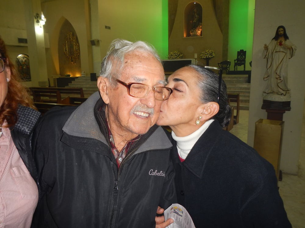 A woman affectionately kisses an elderly man on the cheek indoors.