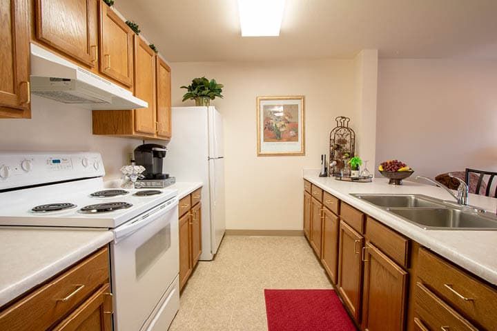 A cozy kitchen with wooden cabinets and white appliances, featuring a red rug and decorative elements.