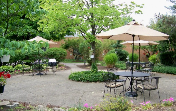 A serene garden patio with tables, chairs, and umbrellas surrounded by lush greenery.
