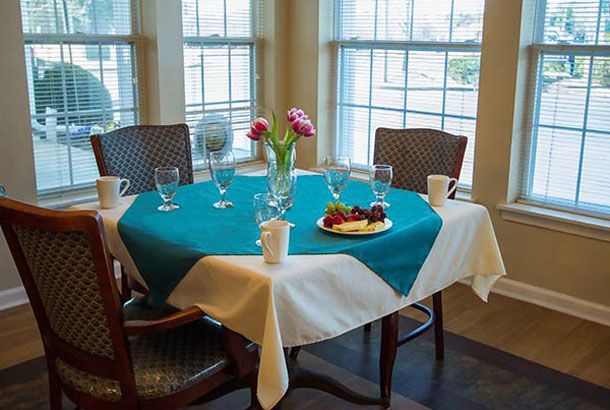 A cozy dining table set with a teal and white tablecloth, adorned with flowers, glasses, and coffee cups by the window.