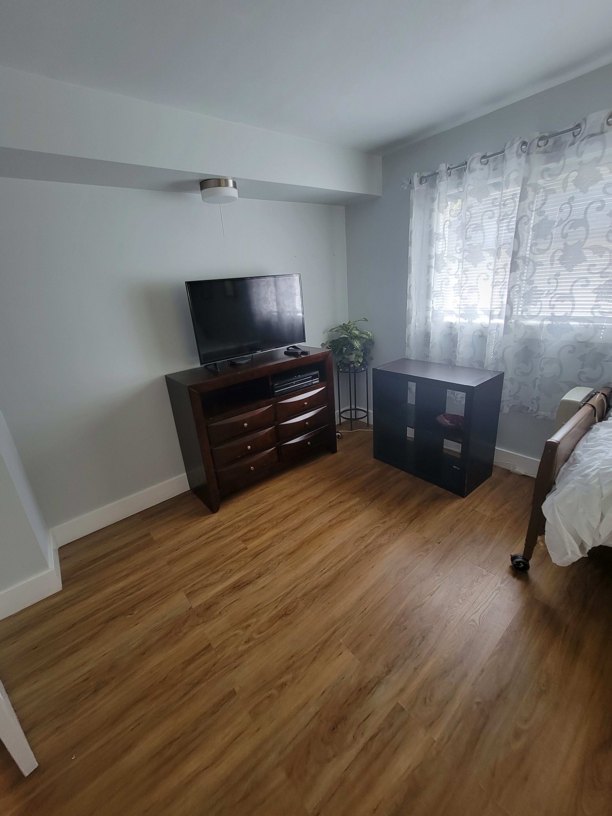 A cozy bedroom with a TV on a dresser, hardwood floors, and curtains by the window.