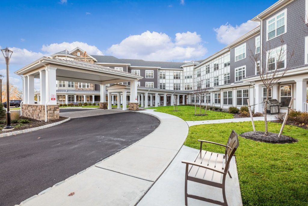 A modern three-story building with a circular driveway and an outdoor bench under a partly cloudy sky.