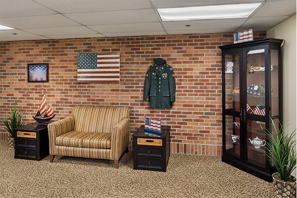 A room with veteran memorabilia, featuring an American flag, a military jacket on a brick wall, and a comfortable armchair.