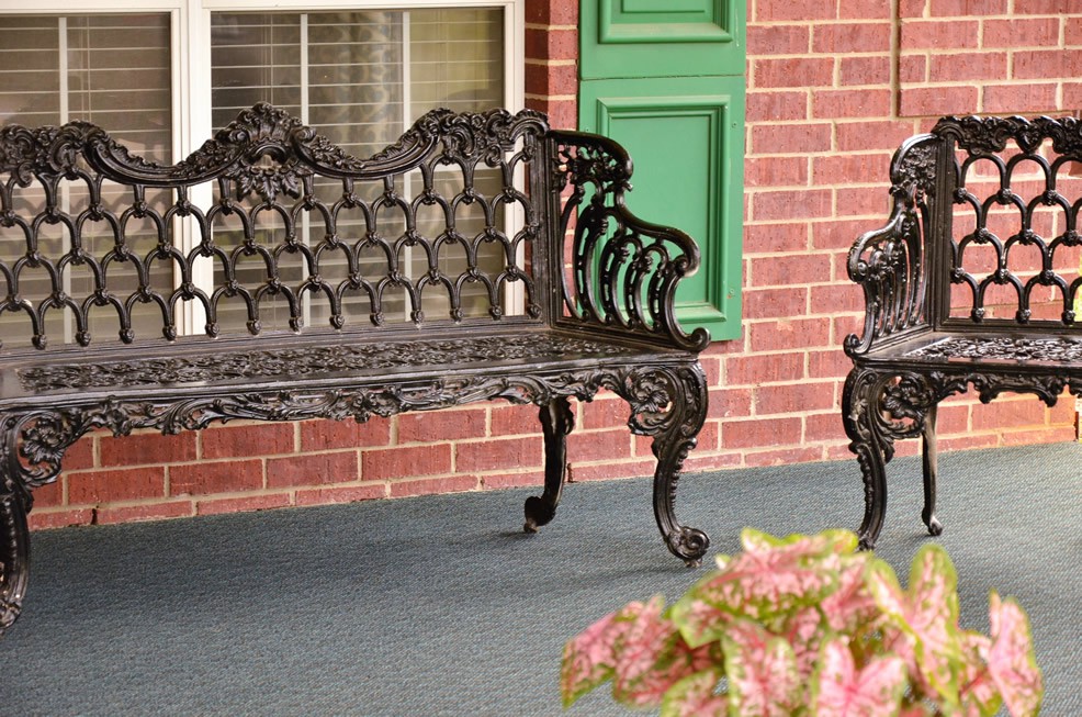 A decorative wrought iron bench and chair on a brick patio with green shutters.