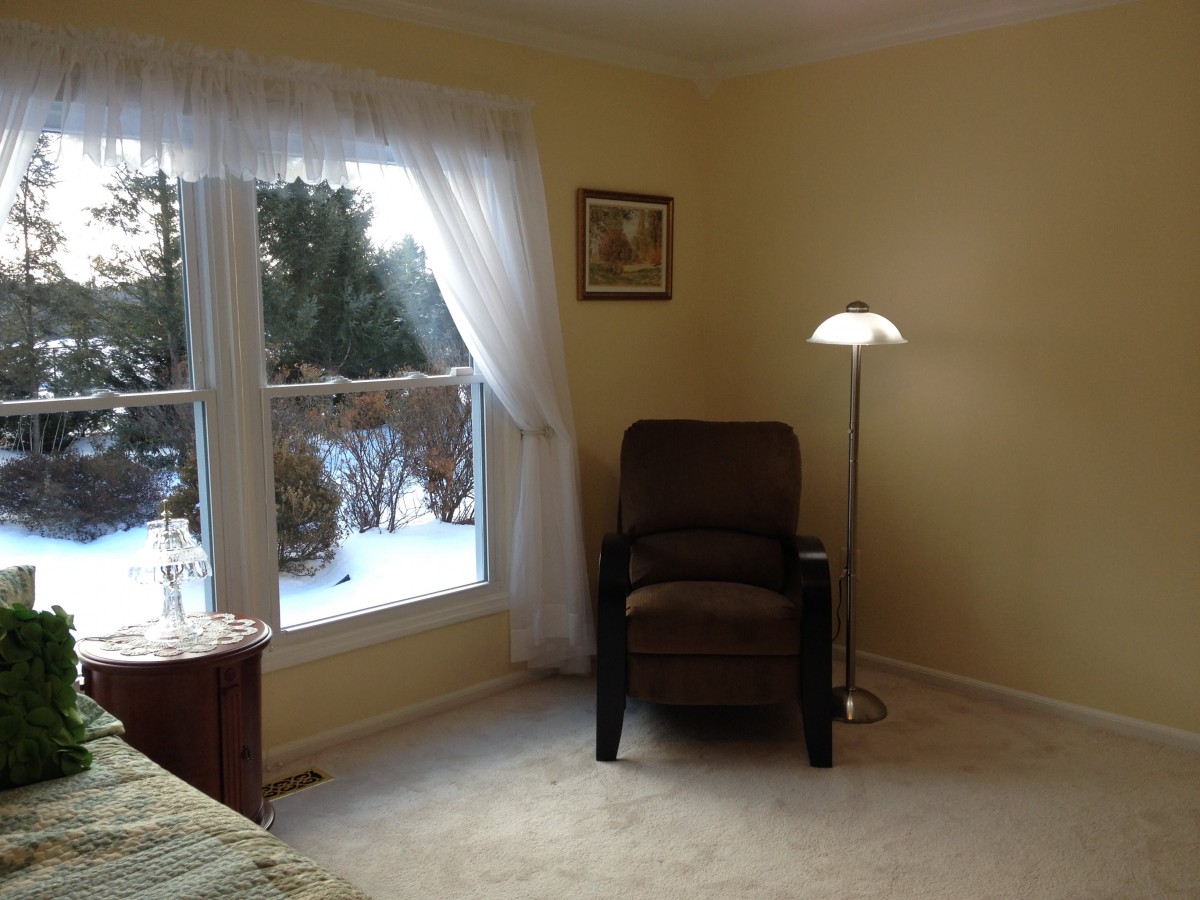 A cozy corner in a living room featuring a comfortable chair next to a tall lamp, with natural light coming through a curtained window overlooking a snowy landscape.